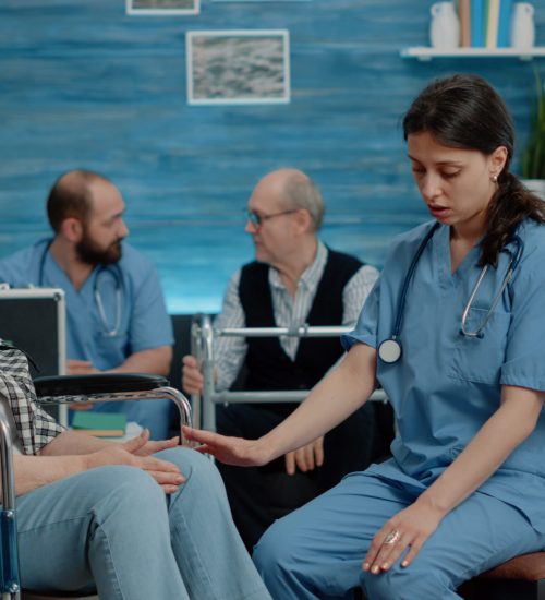 Nurse consulting retired woman sitting in wheelchair at nursing home clinic. Medical assistant doing healthcare checkup with disabled old patient for treatment and recovery from pain.
